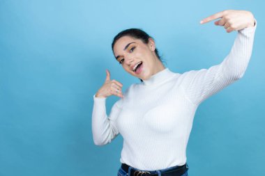 Mavi arka plan üzerine beyaz kazak giyen genç beyaz kadın bana elleriyle jest yapıyor.