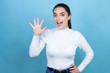 Mavi arka planda beyaz süveter giyen genç beyaz kadın beş numaralı parmaklarıyla işaret ederken kendinden emin ve mutlu gülümsüyor.