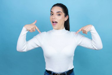 Mavi arka planda beyaz süveter giyen, kendine güvenen, kendini gururlu ve mutlu gösteren genç beyaz bir kadın..