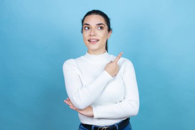 Mavi arka planda beyaz süveter giyen genç beyaz kadın kafası karışmış ve elini ve parmağını yana uzatmış.