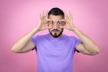 Pembe arka plan üzerine gündelik tişört giyen genç yakışıklı adam parmaklarıyla gözlerini açmaya çalışıyor, uykulu ve yorgun sabah yorgunluğu için.