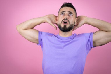 Pembe arka planda rahat bir tişört giyen, kollarını ve ellerini başının ve boynunun arkasına koyup gülümseyen yakışıklı bir adam.