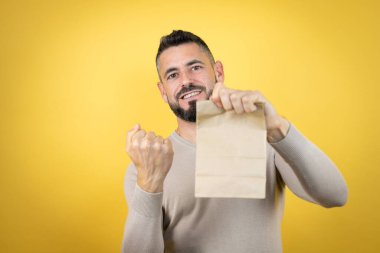 Sarı arka planda sakallı yakışıklı bir adam elinde bir kese kağıdıyla çok mutlu ve heyecanlı bir şekilde kollarını kaldırıp, gülümseyerek ve başarı için bağırarak kazanan bir jest yapıyor.