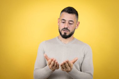 Sarı arka planda süveter giyen sakallı yakışıklı bir adam avuçlarını birbirine değdirip gülümsüyor ya da el hareketi yapıyor. Tutun ve koruyun