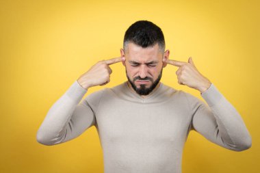 Sakallı yakışıklı bir adam sarı arka planda kazak giyiyor. Yüksek sesli müzik sesi için kulaklarını parmaklarıyla kapatıyor. Sağır kavramı.