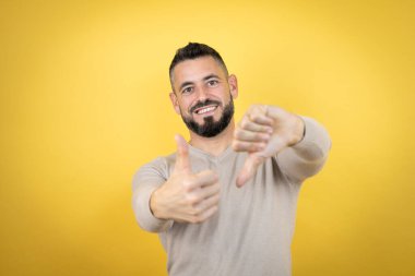 Sarı arka plan üzerine kazak giyen sakallı yakışıklı bir adam baş parmak kaldırıyor, anlaşmazlık ve anlaşma ifadesi takınıyor. Çılgın çatışma