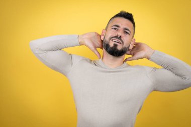 Sakallı yakışıklı adam sarı arka planda süveter giyiyor rahatlıyor ve esniyor, kolları ve elleri başının arkasında mutlu bir şekilde gülümsüyor.