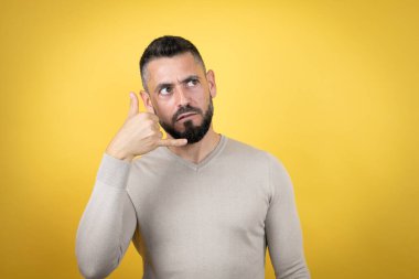 Sarı arka plan üzerine kazak giyen sakallı yakışıklı adam telefon hareketlerini elle ve parmaklarla yapıyor telefonda konuşuyor gibi.