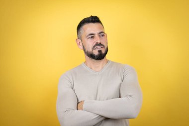 Sakallı, sarı arka planda süveter giyen yakışıklı adam yorgun ve kollarını kavuşturmaktan sıkılmış görünüyor.