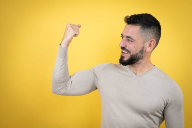 Sarı arka planda süveter giyen sakallı yakışıklı adam gururla gülümseyen kol kaslarını gösteriyor.