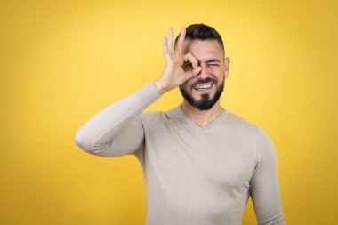 Sakallı, sarı arka planda süveter giyen yakışıklı adam gülümseyen yüzüyle şok olmuş, parmaklarının arasından bakan.