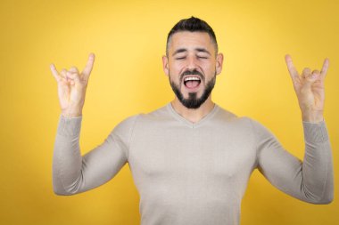 Sarı arka planda süveter giyen yakışıklı adam çılgın bir ifadeyle el kaldırarak rock sembolü yapıyor.
