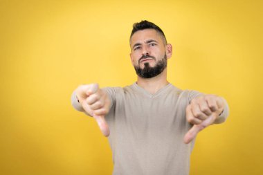 Sakallı, sarı arka planda süveter giyen, kızgın yüzlü, negatif işaretli, baş parmaklı, hoşlanmayan yakışıklı bir adam.