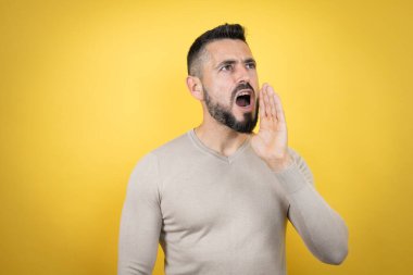 Sakallı, sarı arka planda süveter giyen yakışıklı adam bağırıyor ve ağızdan ağıza bağırıyor.