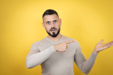 Sarı arka planda süveter giyen yakışıklı bir adam elinde olan bir şeyi gösterip işaret ediyor.