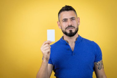 Sarı arka planda mavi polo tişört giyen sakallı yakışıklı adam gülümsüyor ve elinde beyaz kart tutuyor.