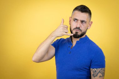 Sarı arka planda mavi polo tişörtlü yakışıklı adam telefon hareketlerini el ve parmaklarıyla yapıyor telefonda konuşuyor gibi.