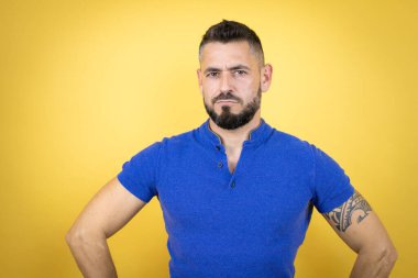 Handsome man with beard wearing blue polo shirt over yellow background skeptic and nervous, disapproving expression on face with arms in waist clipart