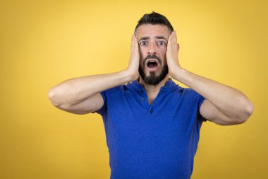 Sakallı, sarı arka planda mavi polo tişörtlü yakışıklı adam çılgın ve elleri kafasında korkmuş, şoktan korkmuş ve şaşırmış.