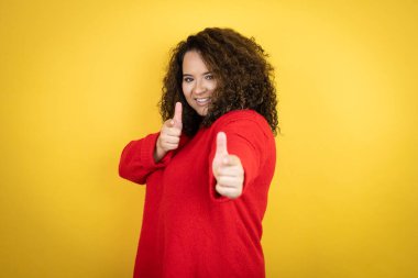 Sarı arka plan üzerine kırmızı süveter giyen genç Afro-Amerikalı kadın seni ve kamerayı parmaklarıyla gösteriyor, gülümsüyor ve neşeli.