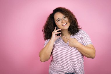 Pembe arka planda kırmızı çizgili tişört giyen Afro-Amerikalı genç bir kadın akıllı telefondan konuşurken mutlu bir şekilde el ve parmak göstererek gülümsüyor.