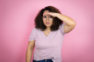 Genç Afro-Amerikalı kadın pembe arka planda kırmızı çizgili tişört giyiyor çok mutlu ve gülümsüyor başlarını kaldırıp uzaklara bakıyor. arama kavramı.