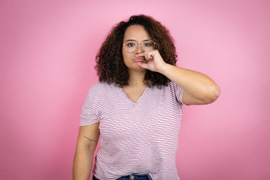 Genç Afro-Amerikalı kadın pembe arka plan ağzının üzerine kırmızı çizgili tişört giyer ve dudakları parmaklarıyla kapanır. Gizli ve sessiz, tabu konuşması