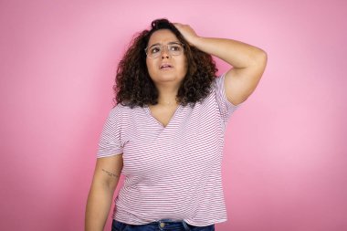 Pembe arka planda kırmızı çizgili tişört giyen Afro-Amerikalı genç bir kadın bir elini kafasına koyup sanki bir şey unutmuş gibi gülümsüyordu.