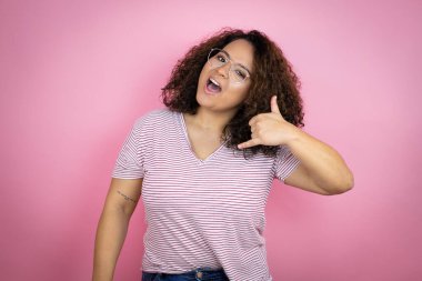 Pembe arka planda kırmızı çizgili tişört giyen genç Afro-Amerikalı kadın telefonla el ve parmak hareketleriyle gülümsüyor telefonda konuşuyor gibi.