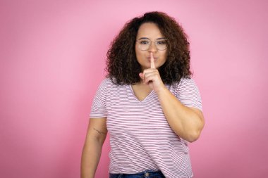 Pembe arka planda kırmızı çizgili tişört giyen genç Afro-Amerikan kadın sessiz olup dudaklarını öpmek istiyor. Sessizlik ve gizli konsept.