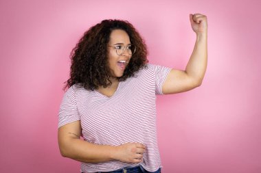 Pembe arka planda kırmızı çizgili tişört giyen Afro-Amerikalı genç bir kadın gururla gülümseyen kol kaslarını gösteriyor.