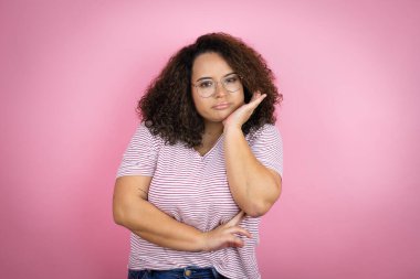 Pembe arka planda kırmızı çizgili tişört giyen genç Afrikalı Amerikalı kadın çapraz kollardan sıkılmış ve yorgun göründüğünü düşünüyor.