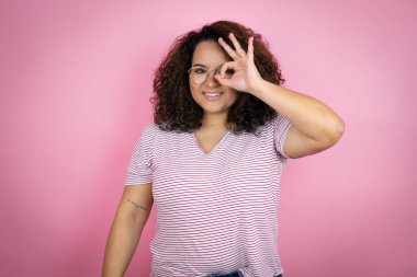 Pembe arka planda kırmızı çizgili tişört giyen Afro-Amerikalı genç bir kadın gülümseyen yüz ve parmaklarından bakan gözlerle şok olmuş bir jest yapıyor.