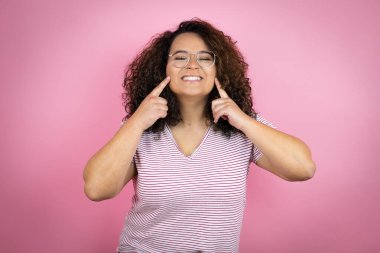 Pembe arka planda kırmızı çizgili tişört giyen genç Afro-Amerikalı kadın gülümsüyor ve parmaklarıyla ve ağzıyla işaret ediyor.