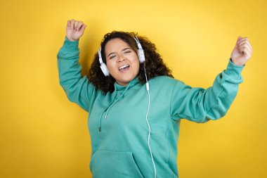Genç Afro-Amerikalı kız spor kıyafetleri giyip kulaklık takıyor, dans ediyor ve mutlu bir yüzle müzik dinliyor. Kendine güvenen bir gülümsemeyle dişlerini gösteriyor.