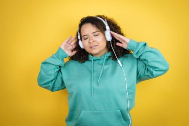 Genç Afro-Amerikalı kız spor kıyafetleri giyip kulaklık takıyor, dans ediyor ve mutlu bir yüzle müzik dinliyor. Kendine güvenen bir gülümsemeyle dişlerini gösteriyor.