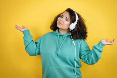 Spor kıyafetleri giyen, kulaklık takan, kolları ve elleri havada, kafası karışmış genç Afrikalı Amerikalı kız.