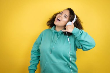 Genç Afrikalı Amerikalı kız spor kıyafetleri giyiyor ve kulaklık takıyor. Gülümsüyor. El ve parmaklarıyla telefon hareketi yapıyor.