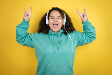 Genç Afrikalı Amerikalı kız spor kıyafetleri giyiyor ve kulaklık takıyor çılgın bir ifadeyle bağırıyor elleri havada rock sembolü yapıyor.