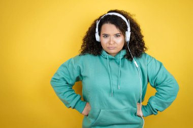 Genç Afrikalı Amerikalı kız spor kıyafetleri giyiyor ve kulaklık takıyor. Kuşkucu ve gergin. Yüzünde kolları belinde tasvip etmeyen bir ifade var.