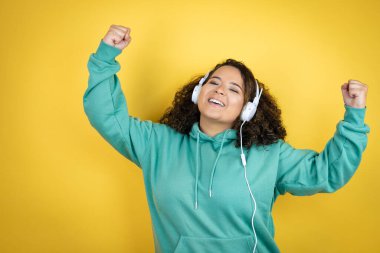 Genç Afrikalı Amerikalı kız spor kıyafetleri giyiyor ve kulaklık takıyor. Çok mutlu ve heyecanlı. Kollarını kaldırıp, gülümseyerek ve başarı için bağırarak kazanan jestler yapıyor..