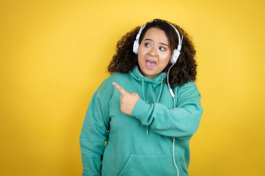 Genç Afrikalı Amerikalı kız spor kıyafetleri giyiyor ve kulaklık takıyor gülümsüyor ve parmaklarını yana doğru uzatıyor.