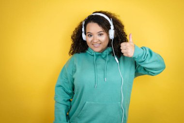 Genç Afrikalı Amerikalı kız spor kıyafetleri giyiyor ve kulaklık takıyor. Başparmağıyla gülümsüyor ve mutlu bir şekilde olumlu jest yapıyor. Neşeli ifade
