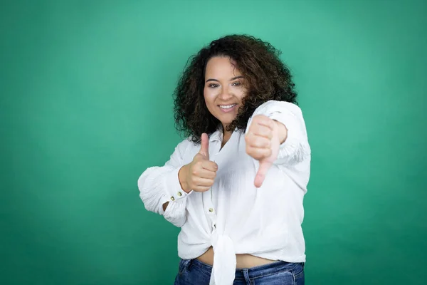 Young african american girl wearing white shirt over green background Doing thumbs up and down, disagreement and agreement expression. Crazy conflict