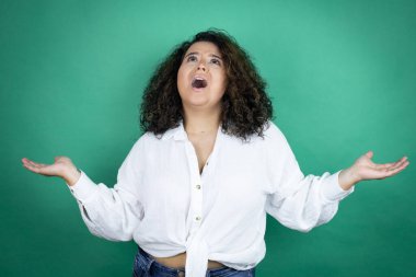 Yeşil arka planda beyaz gömlek giyen, elleri ve kolları havada, kafası karışmış genç Afrikalı Amerikalı kız.