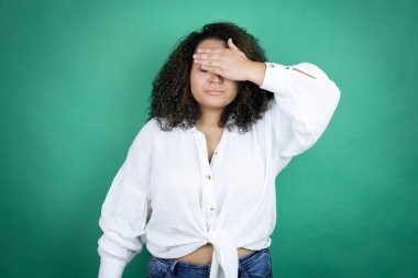 Genç Afrikalı Amerikalı kız yeşil arka planda beyaz gömlek giyiyor ciddi ve elleriyle gözlerini kapatıyor.
