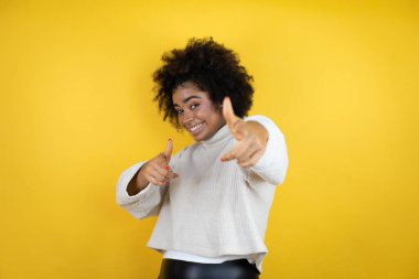 Sarı arka plan üzerine günlük süveter giyen Afro-Amerikalı kadın seni ve kamerayı işaret ediyor, parmaklarıyla gülümsüyor ve neşeli.