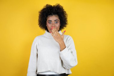 Sarı arka plan üzerine gündelik süveter giyen Afrikalı Amerikalı kadın ağızlarını eliyle kapatıyor, şok geçiriyor ve hata yapmaktan korkuyor. şaşırmış ifade