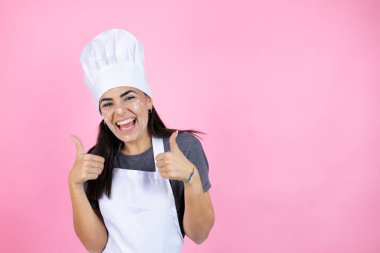Genç İspanyol kadın fırıncı üniforması giyiyor. Pembe arka plan işareti üzerinde un var. Elleriyle olumlu jest yapıyor. Neşeli ifade ve kazanan jest.