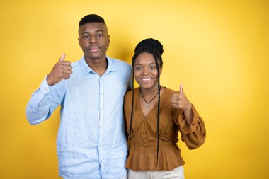 Sarı arka planda duran genç Afro-Amerikan çifti elleriyle olumlu bir jest yapıyor, gülümsüyor ve mutlu. Neşeli ifade